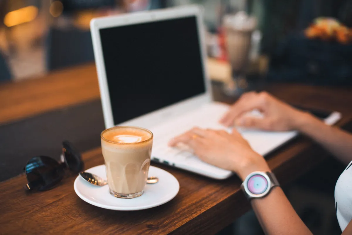 Person typing on a laptop next to a latte on a wooden table